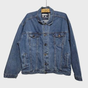 May include: A blue denim jacket featuring a classic collar, long sleeves, and a front button closure. The garment includes two chest pockets with buttoned flaps and two side pockets. A white label with the text LK9 is visible at the neck.