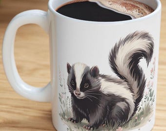 May include: White ceramic coffee mug with a black and white illustration of a skunk. The skunk is facing the left side of the mug and has a black nose and eyes. The skunk is surrounded by green grass and pink flowers.