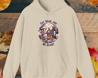 Puede incluir: Una sudadera con capucha de color beige que presenta un gráfico circular en el pecho. La ilustración muestra a una bruja, un esqueleto y un hombre lobo alrededor de una mesa con calabazas y dulces. El texto dice EAT, DRINK, AND BE SCARY.