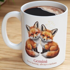 Op de afbeelding: Witte keramische mok met twee oranje vossen met zwarte oren. De mok heeft de tekst "Gemini Never boring, always curious" en is gevuld met donkere koffie.