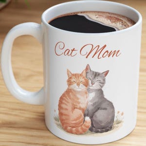 May include: White ceramic coffee mug with a brown handle. The mug has a black coffee inside and a design of two cats cuddling with the text "Cat Mom" in brown lettering.