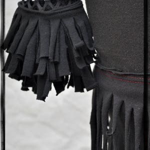 May include: Black shirt with a fringe trim around the bottom and sleeves. The fringe is made of fabric strips that are cut and frayed. The shirt has a mesh panel on the upper part of the sleeves.