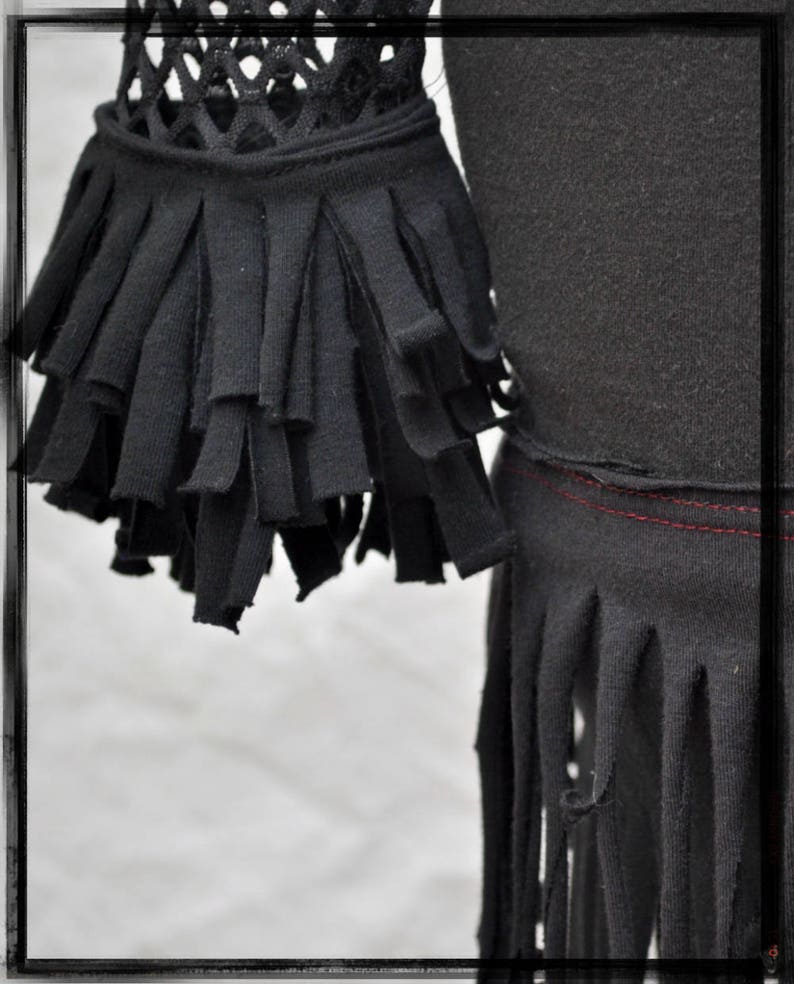 May include: Black shirt with a fringe trim around the bottom and sleeves. The fringe is made of fabric strips that are cut and frayed. The shirt has a mesh panel on the upper part of the sleeves.