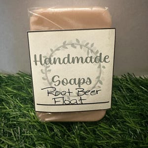 Puede incluir: Una pastilla de jabón artesanal envuelta en plástico transparente. El jabón es de color marrón claro y tiene una etiqueta que dice "Handmade Soaps Root Beer Float".
