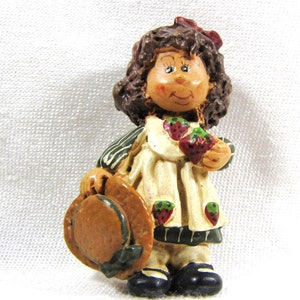 Peut inclure: Une figurine représentant une fille aux cheveux bruns portant une robe blanche à rayures vertes et noires, tenant des fraises. Elle porte un chapeau brun et a une expression douce sur son visage.