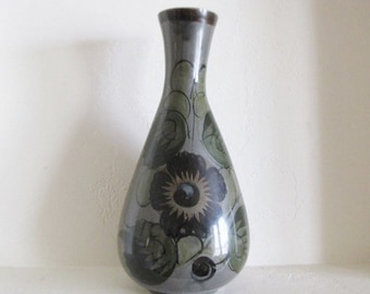 May include: A gray ceramic vase with a dark floral pattern. The vase has a teardrop shape with a narrow neck and a brown rim. The flowers are dark with green leaves.
