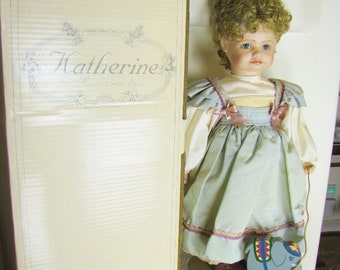 May include: A porcelain doll named "Katherine" in a box, wearing a light blue dress with pink accents. A wooden elephant pull toy with red wheels sits nearby. The doll has curly blonde hair and blue eyes.