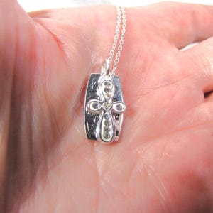 Puede incluir: Collar de plata con un colgante de cruz adornado con brillantes transparentes. El colgante tiene las palabras "We are family" grabadas.