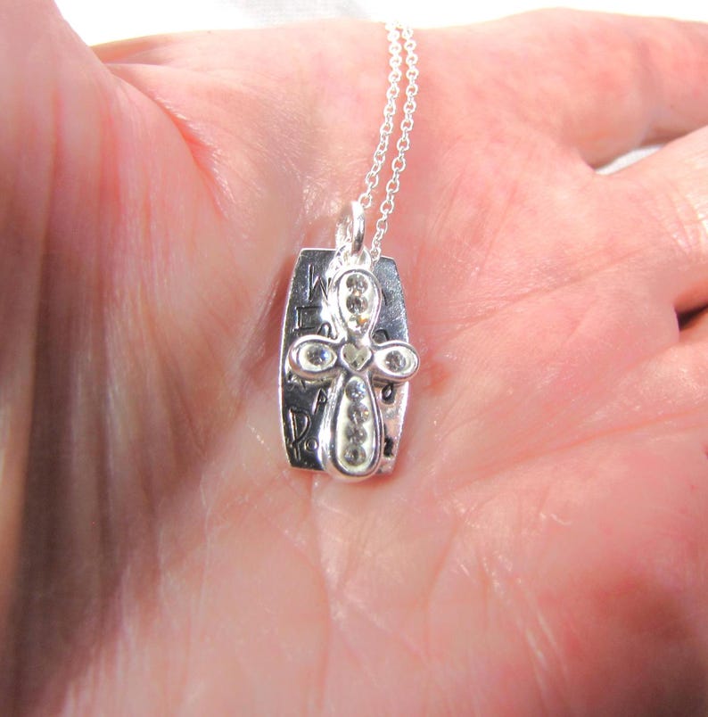 Puede incluir: Collar de plata con un colgante de cruz adornado con brillantes transparentes. El colgante tiene las palabras "We are family" grabadas.