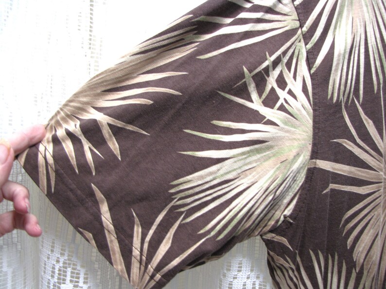 May include: Close-up of a brown short-sleeved shirt with a palm leaf pattern in shades of tan and green. The fabric is visible, showing the texture and the stitching of the sleeve.