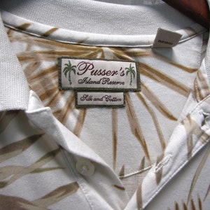 May include: Close-up of a white collared shirt with a brown and tan leaf pattern. The shirt features a label that reads "Pusser's Island Reserve Silk and Cotton". The shirt is hanging on a wooden hanger.