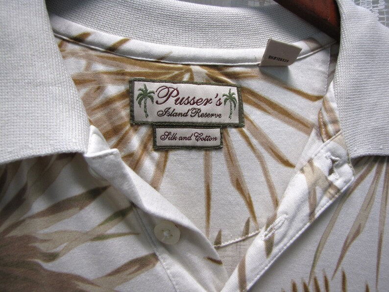 May include: Close-up of a white collared shirt with a brown and tan leaf pattern. The shirt features a label that reads "Pusser's Island Reserve Silk and Cotton". The shirt is hanging on a wooden hanger.