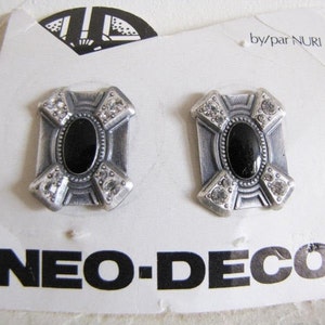 May include: A pair of silver earrings with black oval stones and clear rhinestones. The earrings are in the shape of a cross and have the text "NEO-DECO" on the card. by/par NURI