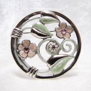 May include: A silver tone brooch with a circular design featuring pink flowers and green leaves. The brooch has a white enamel background.