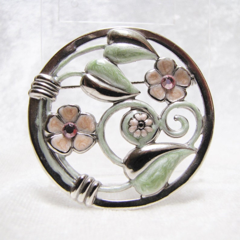 May include: A silver tone brooch with a circular design featuring pink flowers and green leaves. The brooch has a white enamel background.
