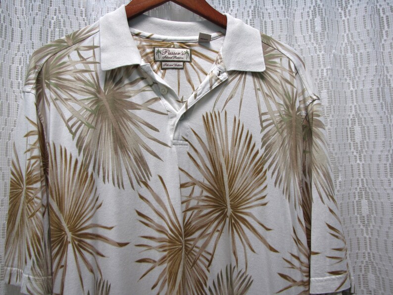 May include: White polo shirt with a tropical palm leaf pattern in shades of brown and green. The shirt has a light-colored collar and a button-down placket. The brand name "Pusser's Island Marine" is visible on a tag.