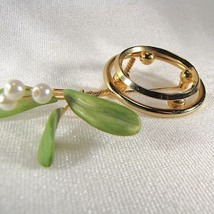 May include: A gold-toned brooch with a double-ring design, accented with small gold spheres. The brooch is paired with a sprig of green leaves and faux pearls, creating a nature-inspired jewelry piece.