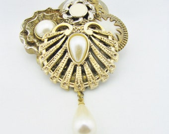 May include: A vintage gold-tone brooch with a seashell design, featuring a large pear-shaped faux pearl pendant. The brooch has intricate detailing and is adorned with additional faux pearls and decorative elements, creating a classic, elegant look.