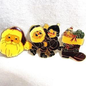 Puede incluir: Tres broches navideños de esmalte. Uno presenta a Papá Noel con barba amarilla y gorro rojo. Otro muestra a dos niños abrazándose. El tercero es un calcetín navideño con regalos.