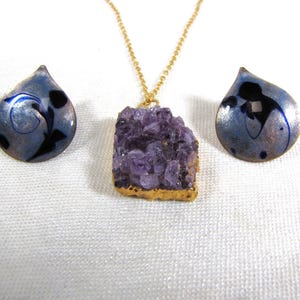 May include: A set of amethyst jewelry featuring a large, raw amethyst pendant on a gold chain and two matching earrings. The earrings are teardrop shaped with a blue and silver enamel design.