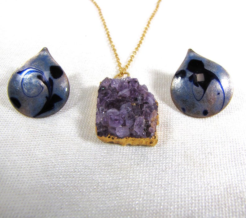 May include: A set of amethyst jewelry featuring a large, raw amethyst pendant on a gold chain and two matching earrings. The earrings are teardrop shaped with a blue and silver enamel design.