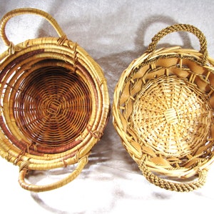 May include: Two round woven baskets with handles. One basket is primarily light brown with a darker brown interior. The other basket is light tan with a braided handle and leather accents. Both baskets are empty.