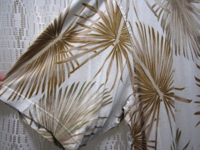 May include: White short-sleeved shirt with a brown and green palm leaf pattern. The shirt's fabric is visible, showing the texture and the stitching of the sleeve.