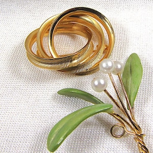 May include: A gold-toned brooch with three interlocking rings and a textured surface. Beside it is a gold-wired sprig with green leaves and three faux pearls. The brooch and sprig are set against a white background.
