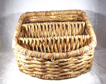 May include: A woven basket with a rectangular shape and a divided interior. The basket is made of natural-colored, tightly woven fibers, showcasing a rustic aesthetic. The basket's design includes a curved front and a sturdy base, suitable for storage.