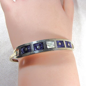 May include: Silver-tone hinged bracelet with a rectangular design. The bracelet features alternating dark purple and white mother-of-pearl inlays. Each purple inlay has a small silver dot in the center. The bracelet is on a wrist.