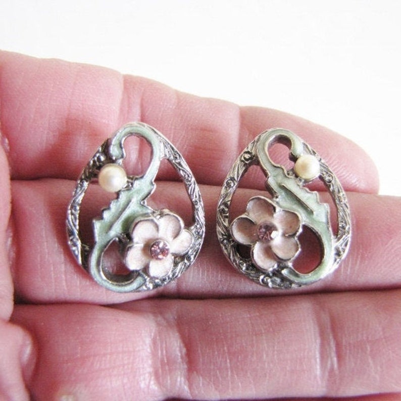 May include: A pair of silver tone earrings with a pink flower and a pearl accent. The earrings are in the shape of an oval and have a delicate, intricate design.