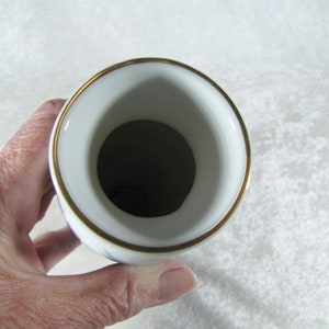 May include: White porcelain vase with a gold rim and blue floral pattern. The vase is empty and the inside is visible.