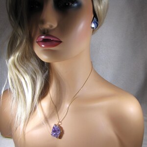 May include: A gold chain necklace with a large, purple amethyst crystal pendant. The pendant is a natural, raw crystal with a rough, irregular shape.