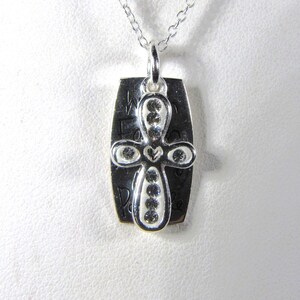 Puede incluir: Collar de plata con un colgante de cruz con un coraz&oacute;n y piedras transparentes. El colgante tiene las palabras "We Found Peace" grabadas.