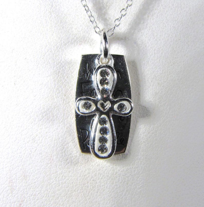 Puede incluir: Collar de plata con un colgante de cruz con un coraz&oacute;n y piedras transparentes. El colgante tiene las palabras "We Found Peace" grabadas.