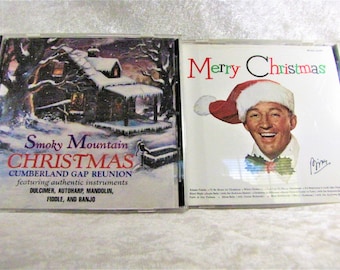 May include: Two Christmas music CDs. One features a snowy cabin scene with "Smoky Mountain Christmas" text. The other CD cover has a man wearing a Santa hat with "Merry Christmas" text. Both are in clear plastic cases.