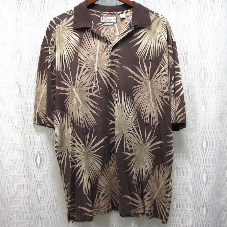 May include: Brown short-sleeved polo shirt with a palm leaf pattern in shades of tan and brown. The shirt has a collared neckline and a button-up placket. The shirt is made of a lightweight fabric.