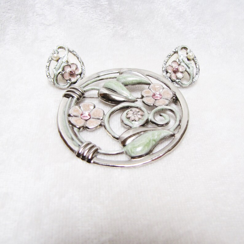 May include: A silver tone brooch and earrings set with a floral design. The brooch is round and features pink and green enamel flowers with rhinestones in the center. The earrings are small and feature a single pink enamel flower with a rhinestone in the center.
