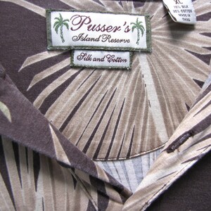May include: Close-up of a brown silk and cotton shirt with a palm leaf pattern. The shirt features a collar and buttons. The label reads "Pusser's Island Reserve".