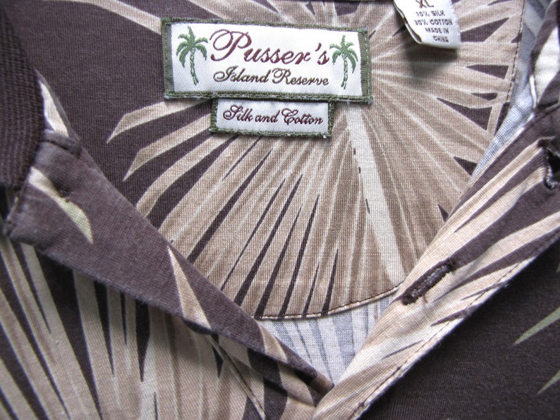 May include: Close-up of a brown silk and cotton shirt with a palm leaf pattern. The shirt features a collar and buttons. The label reads "Pusser's Island Reserve".