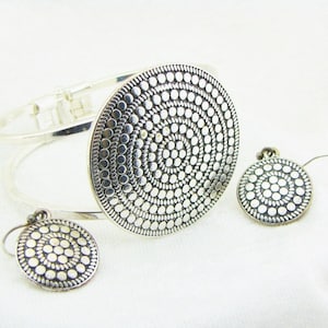 Puede incluir: Un brazalete de plata con un gran colgante circular y pendientes a juego. El colgante y los pendientes tienen un diseño texturizado de círculos concéntricos de pequeños puntos.