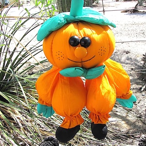 Könnte beinhalten: Eine Kürbisfigur aus Stoff mit einem leuchtend orangefarbenen runden Kopf, schwarzen Knopfaugen, einer kleinen orangefarbenen Nase und einem aufgestickten Lächeln. Sie trägt einen türkisfarbenen Hut und eine Fliege sowie bauschige Ärmel und schwarze Füße.