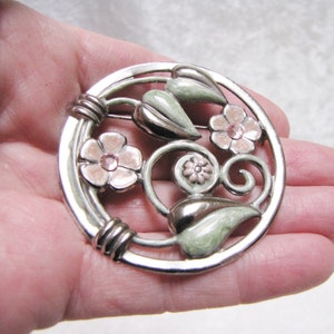 May include: A silver tone brooch with a circular design featuring pink flowers with rhinestones and green leaves. The brooch has a detailed, intricate design.
