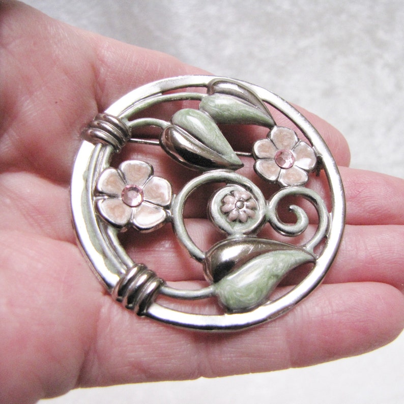 May include: A silver tone brooch with a circular design featuring pink flowers with rhinestones and green leaves. The brooch has a detailed, intricate design.