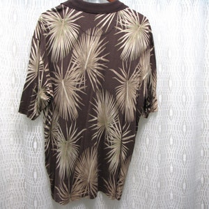 May include: Brown polo shirt with a palm leaf pattern in tan and beige. The shirt has short sleeves and a brown collar. The back of the shirt is visible, showcasing the all-over tropical design.