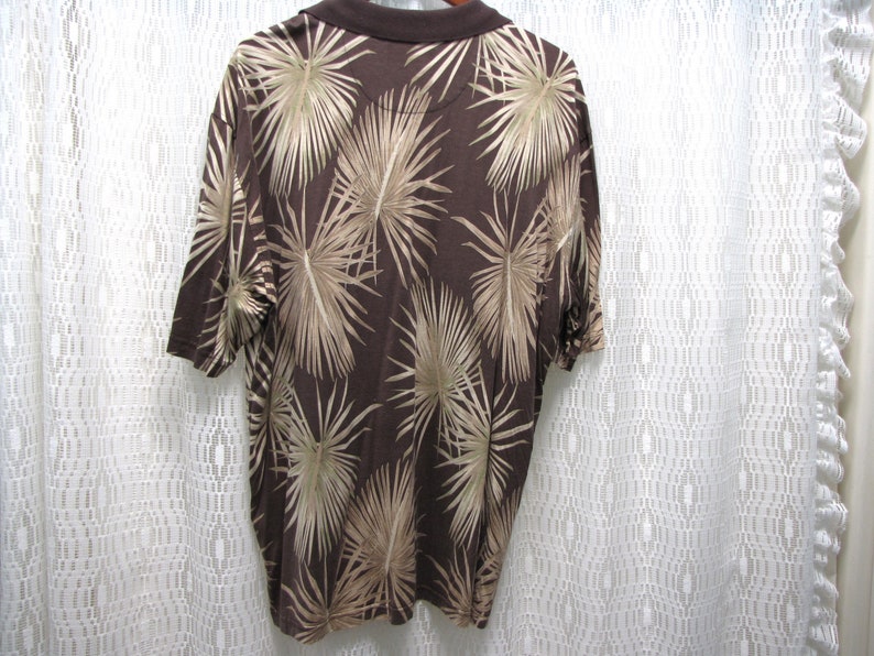 May include: Brown polo shirt with a palm leaf pattern in tan and beige. The shirt has short sleeves and a brown collar. The back of the shirt is visible, showcasing the all-over tropical design.