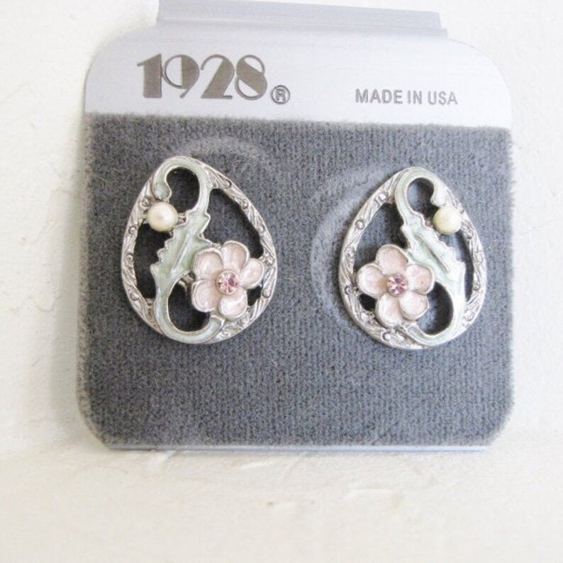 May include: A pair of silver tone earrings with a floral design. The earrings feature a pink flower with a rhinestone center, green leaves, and a white pearl. The earrings are packaged on a gray card with the text "1928" and "Made in USA".