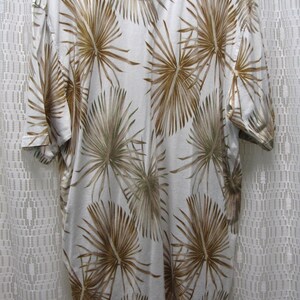 May include: A short-sleeved, collared shirt with a white background and a repeating pattern of brown palm fronds. The shirt is hanging on a hanger, showcasing the back of the garment.
