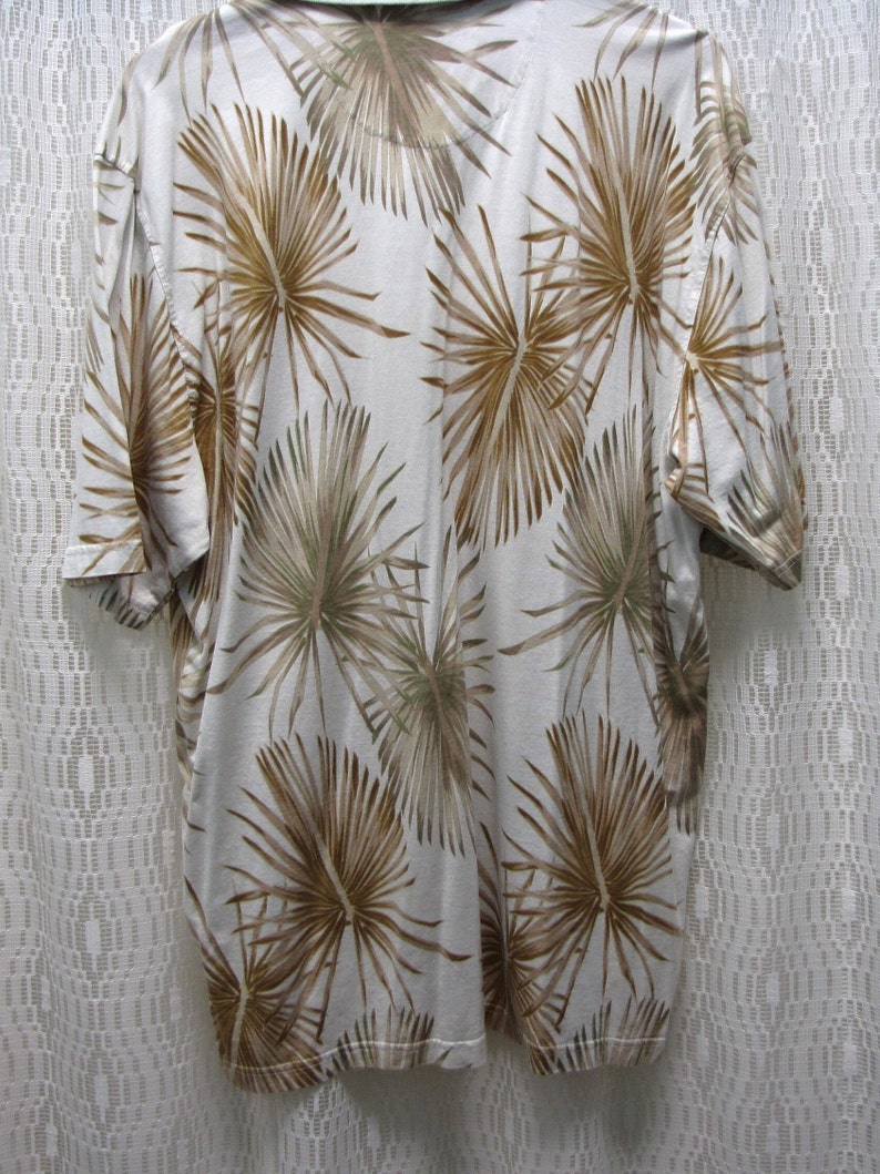 May include: A short-sleeved, collared shirt with a white background and a repeating pattern of brown palm fronds. The shirt is hanging on a hanger, showcasing the back of the garment.