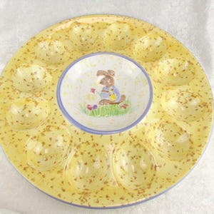 May include: A yellow ceramic deviled egg serving platter with a central dip bowl. The platter has 12 egg indentations and is speckled with brown spots. The dip bowl features a cartoon bunny holding flowers.